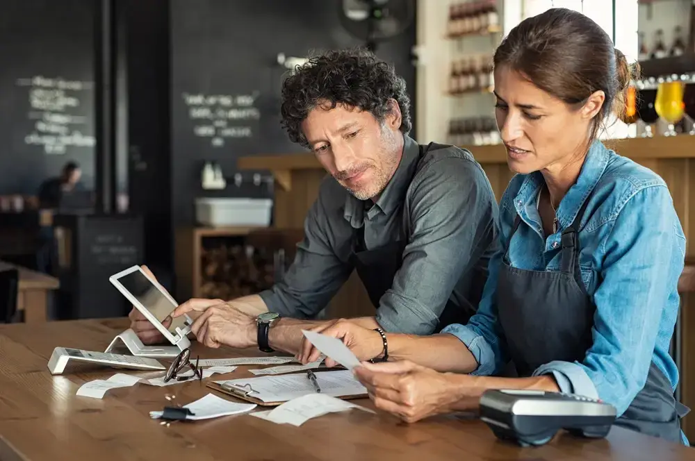 two business owners in a coffee shop, reviewing documents and a digital tablet, possibly discussing their digital marketing consultant's advice on improving their business strategy.