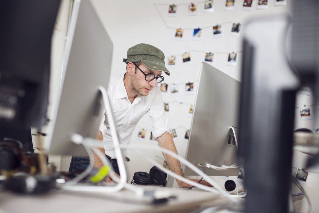 A man wearing a green cap and glasses looking at a computer screen in a modern office. The person appears to be conducting a detailed SEO audit, surrounded by various tech equipment and a wall decorated with pinned photos.