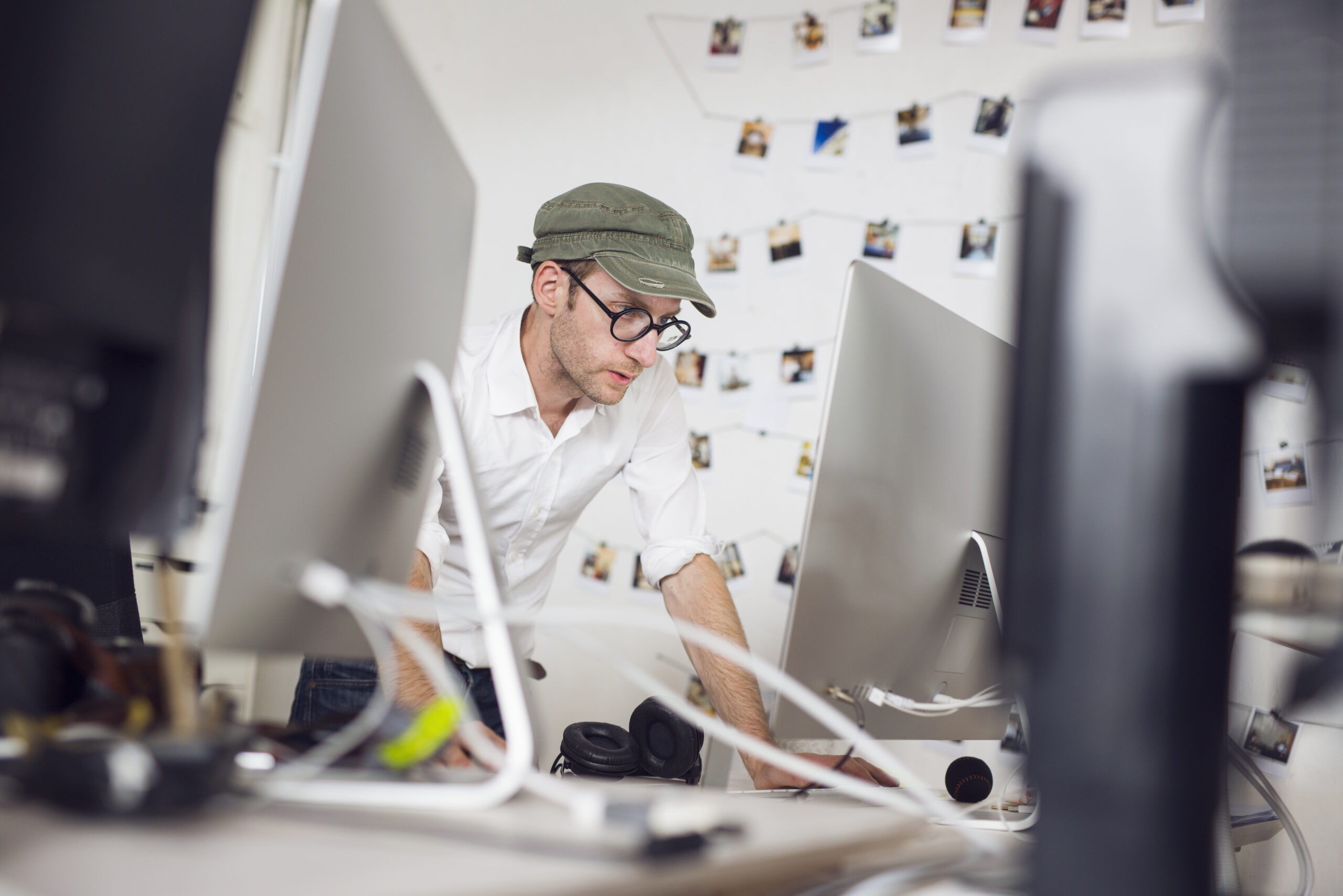 A man wearing a green cap and glasses looking at a computer screen in a modern office. The person appears to be conducting a detailed SEO audit, surrounded by various tech equipment and a wall decorated with pinned photos.
