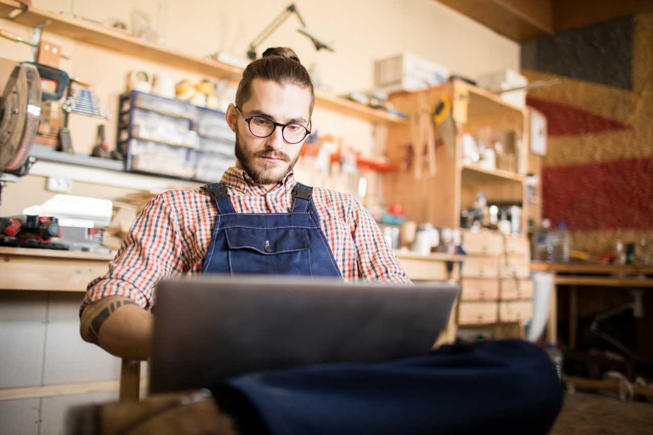 Man in a workshop wearing glasses and overalls, working on a laptop and researching 'Is Google Ads Worth It?' in a woodworking studio, surrounded by tools and materials.