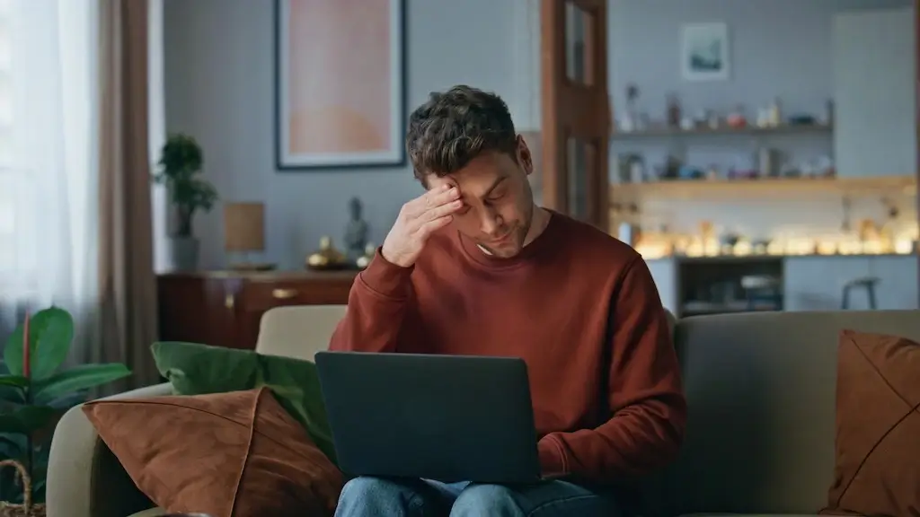 A worried freelancer sitting on a sofa, using a laptop, and holding his forehead in frustration. The image symbolizes the challenges of dealing with SEO mistakes while working remotely.