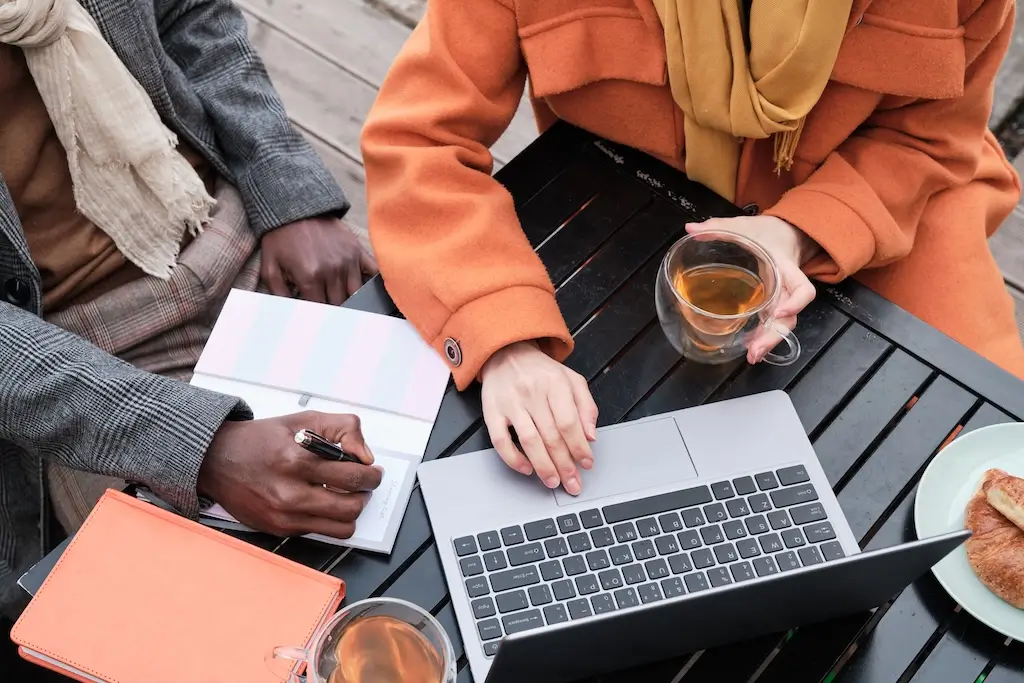 Two people working at an outdoor café, one typing on a laptop and the other taking notes. This setting reflects collaboration, possibly using ChatGPT for keyword research and content planning.