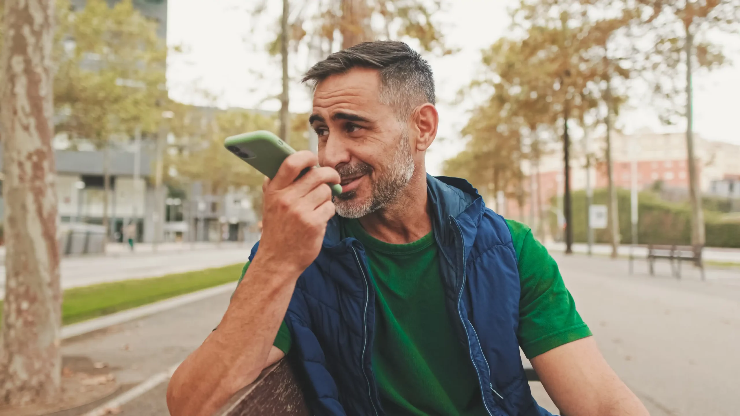 Middle-aged man sitting on a park bench holding a green smartphone near his mouth, appearing to use voice input, symbolizing real-world use of conversational search through voice assistants in casual outdoor settings.